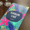 Menu de café imprimé sur Polyart Laser Soft, affichant 'BAOBAB CAFÉ' sur un fond de feuilles et fleurs tropicales colorées, posé sur une table en bois